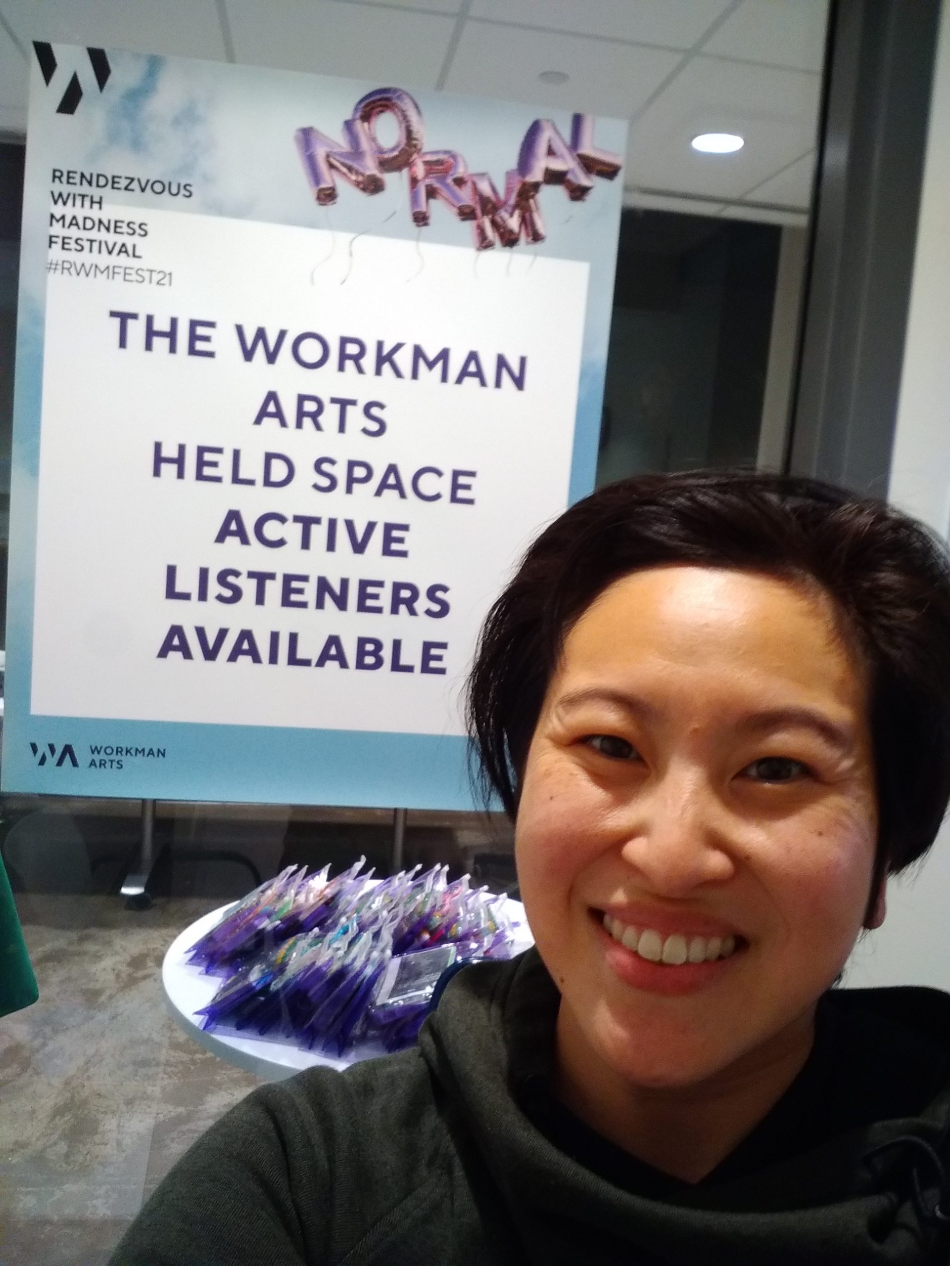 Smiling in front of Workman Arts Held Space Active Listeners Available sign