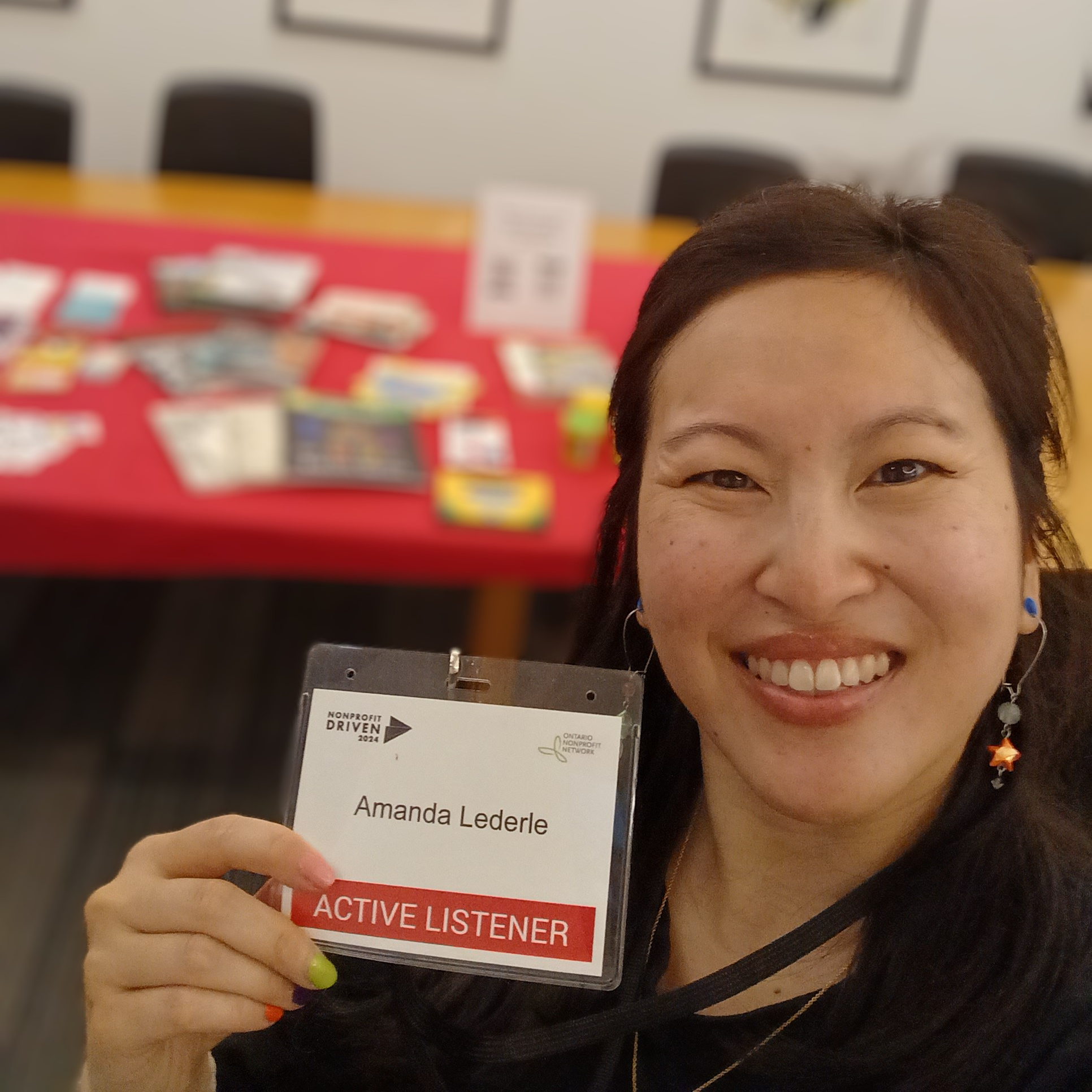 Amanda smiles at the camera while holding their conference badge with their name and Active Listener title. Behind them is a blurred background of their resource table.